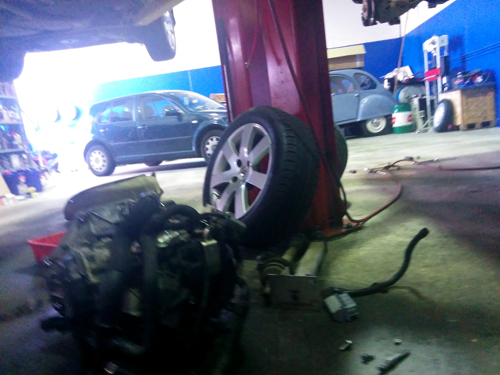 The French Car Care workshop floor: a car up on the two-post hoist, an engine out on the floor, a wheel off, and a grey Citroën 2CV behind